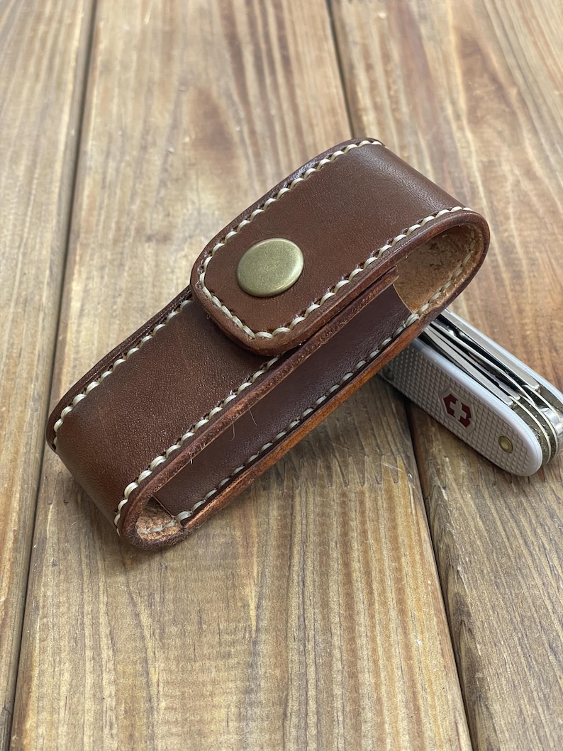 May include: A brown leather knife sheath with white stitching and a brass button closure. A silver and white pocket knife is partially visible next to the sheath. The items are on a wooden surface.