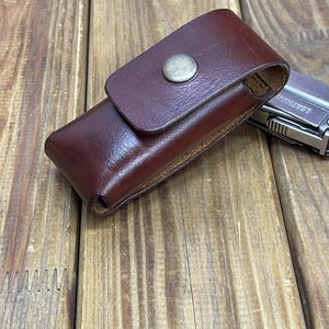 May include: A dark brown leather sheath with a snap closure. The rectangular sheath is designed for a multi-tool. The background is a wooden surface.