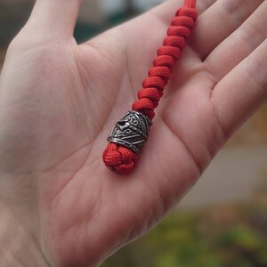 May include: A red paracord lanyard with a silver metal bead. The bead has a detailed design.