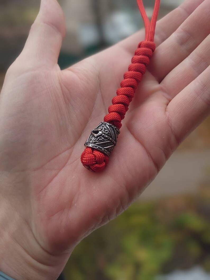 May include: A red paracord lanyard with a silver metal bead. The bead has a detailed design.