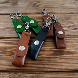 Könnte beinhalten: Fünf Leder-Schlüsselanhänger mit Metallringen und Druckknöpfen. Die Schlüsselanhänger sind in verschiedenen Farben: Braun, Schwarz und Grün.