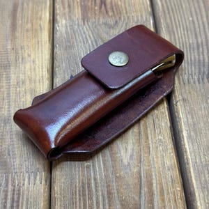 May include: A dark brown leather knife sheath with a brass snap closure. The sheath is designed to hold a folding knife. The leather has a rich, polished appearance, and the design is simple and functional.