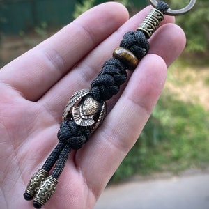 May include: Black paracord keychain with a brass owl charm, a tiger's eye bead, and a brass bead with intricate designs.