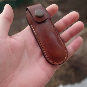 May include: A brown leather knife sheath with a snap closure. The sheath is hand-stitched with a contrasting thread and features a rounded bottom. The leather has a rich, warm tone, and the design is simple and functional.