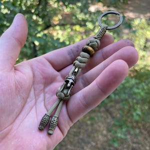 K&ouml;nnte beinhalten: Ein beigefarbener Paracord-Schl&uuml;sselanh&auml;nger mit einer bronzefarbenen Metallperle, die ein r&ouml;misches Helmdesign zeigt. Der Schl&uuml;sselanh&auml;nger enth&auml;lt auch eine Tigerauge-Perle und einen bronzefarbenen Metallring.