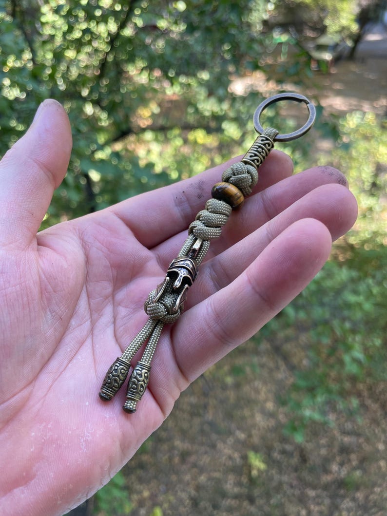 K&ouml;nnte beinhalten: Ein beigefarbener Paracord-Schl&uuml;sselanh&auml;nger mit einer bronzefarbenen Metallperle, die ein r&ouml;misches Helmdesign zeigt. Der Schl&uuml;sselanh&auml;nger enth&auml;lt auch eine Tigerauge-Perle und einen bronzefarbenen Metallring.
