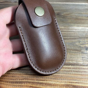 Könnte beinhalten: Ein brauner Leder-Schlüsselanhänger mit Klappenverschluss und einem Messingknopf. Der Schlüsselanhänger hat einen genähten Rand.