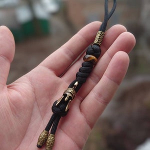 K&ouml;nnte beinhalten: Schwarzer Paracord-Schl&uuml;sselanh&auml;nger mit einem goldfarbenen Helm-Anh&auml;nger, einer Tigerauge-Perle und zwei goldfarbenen Perlen mit filigranen Designs.