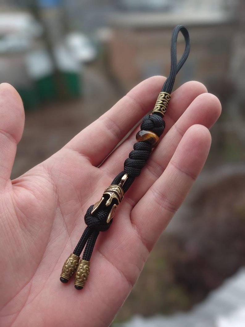 K&ouml;nnte beinhalten: Schwarzer Paracord-Schl&uuml;sselanh&auml;nger mit einem goldfarbenen Helm-Anh&auml;nger, einer Tigerauge-Perle und zwei goldfarbenen Perlen mit filigranen Designs.