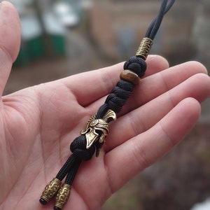 K&ouml;nnte beinhalten: Ein schwarzer Paracord-Schl&uuml;sselanh&auml;nger mit einem goldenen r&ouml;mischen Helm-Anh&auml;nger, einer braunen Tigerauge-Perle und zwei goldenen Perlen.