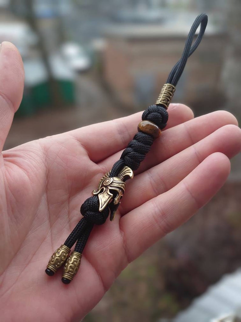 K&ouml;nnte beinhalten: Ein schwarzer Paracord-Schl&uuml;sselanh&auml;nger mit einem goldenen r&ouml;mischen Helm-Anh&auml;nger, einer braunen Tigerauge-Perle und zwei goldenen Perlen.