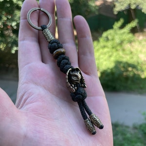 May include: A black paracord keychain with a brass owl charm and a tiger's eye bead. The keychain has a brass ring at the top and two brass end caps.