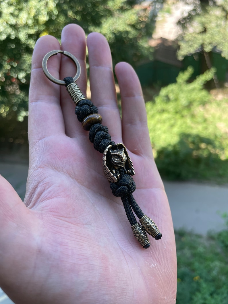 May include: A black paracord keychain with a brass owl charm and a tiger's eye bead. The keychain has a brass ring at the top and two brass end caps.