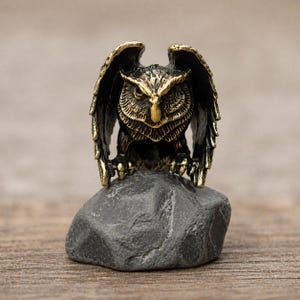 Op de afbeelding: Een bronzen uil beeldje met uitgespreide vleugels, zittend op een grijze steen. De uil heeft gedetailleerde veertjes en een scherpe blik. De achtergrond is een vage houten ondergrond.