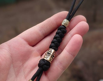 May include: Black paracord keychain with gold metal beads. The beads feature intricate designs, including a cross and a Celtic knot.