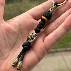 Puede incluir: Un llavero negro y dorado con un llavero de latón. El llavero presenta un dije decorativo de latón, una cuenta de ojo de tigre y detalles decorativos de latón. El llavero está hecho de cordón negro.