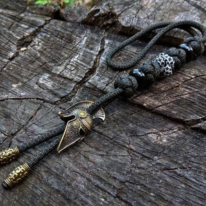 May include: A black and gold bolo tie featuring a bronze-colored Spartan helmet pendant. The tie has a braided black cord with decorative gold-colored tips and a beaded accent. The background is a weathered wooden surface.