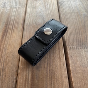 May include: A black leather case with a silver snap closure. The case is rectangular and appears to be stitched along the edges. The case is resting on a wooden surface.