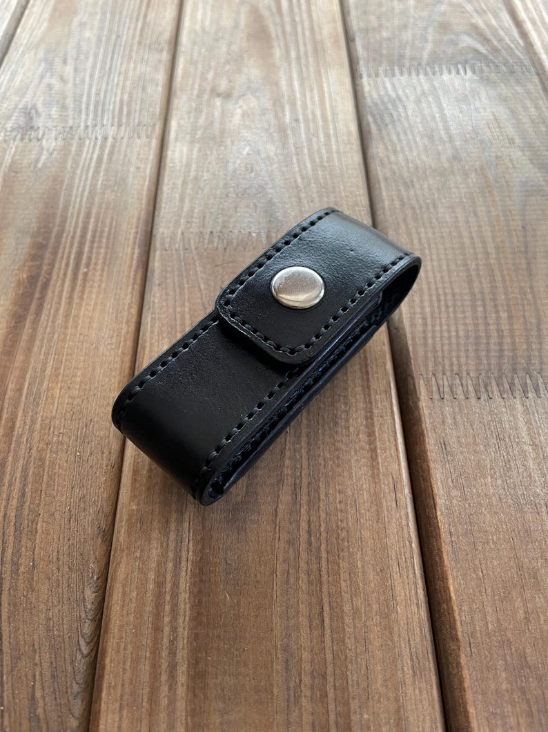 May include: A black leather case with a silver snap closure. The case is rectangular and appears to be stitched along the edges. The case is resting on a wooden surface.