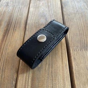May include: A black leather case with a silver snap closure. The case is rectangular and appears to be stitched along the edges. The background is a wooden surface with visible grain.