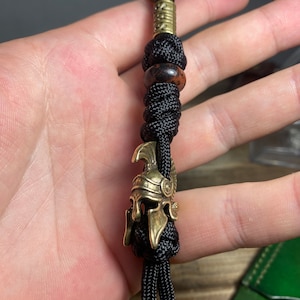 K&ouml;nnte beinhalten: Ein schwarzer Paracord-Schl&uuml;sselanh&auml;nger mit einem r&ouml;mischen Helm-Anh&auml;nger aus Messing, einer braunen Perle und einem Messingring.
