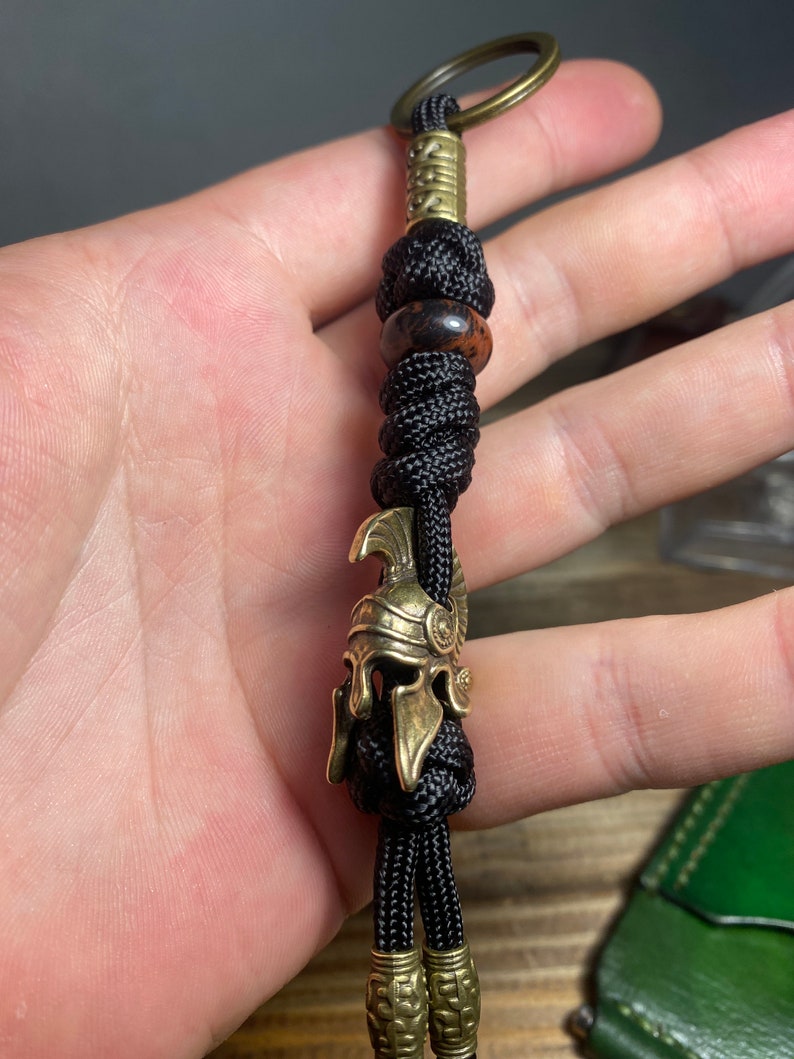 K&ouml;nnte beinhalten: Ein schwarzer Paracord-Schl&uuml;sselanh&auml;nger mit einem r&ouml;mischen Helm-Anh&auml;nger aus Messing, einer braunen Perle und einem Messingring.