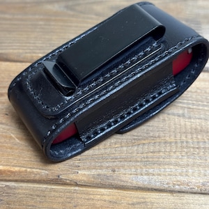 May include: Black leather sheath with a metal clip, designed to hold a small red object. The sheath is stitched with white thread and sits on a wooden surface. The clip is attached to the back of the sheath.