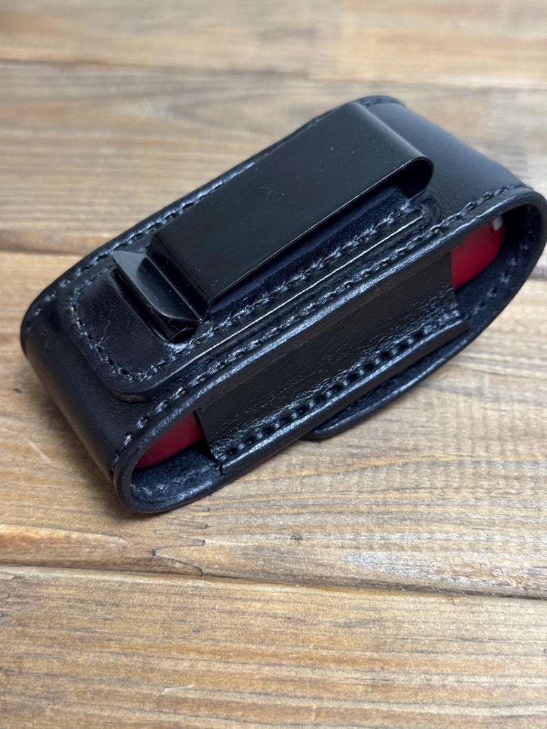 May include: Black leather sheath with a metal clip, designed to hold a small red object. The sheath is stitched with white thread and sits on a wooden surface. The clip is attached to the back of the sheath.