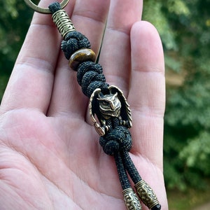 May include: A black paracord keychain with a brass owl charm and a tiger's eye bead. The keychain has two brass beads with intricate designs at the bottom.