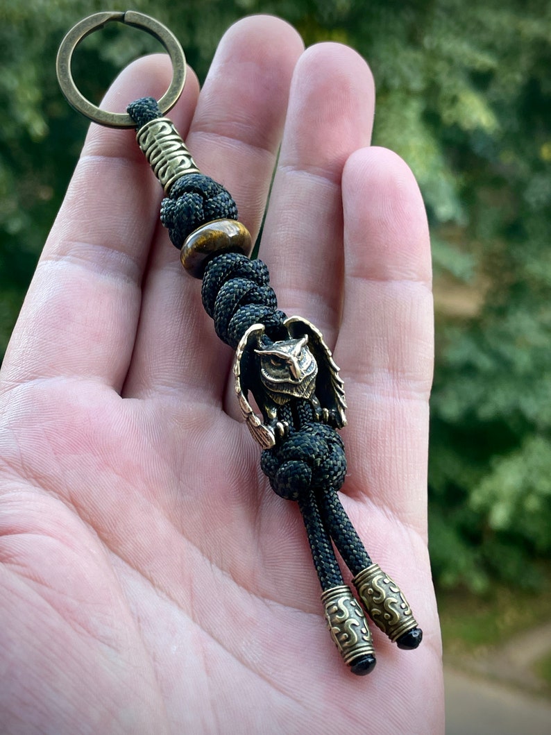 May include: A black paracord keychain with a brass owl charm and a tiger's eye bead. The keychain has two brass beads with intricate designs at the bottom.