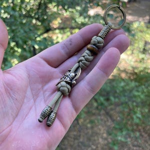 K&ouml;nnte beinhalten: Ein gr&uuml;ner Paracord-Schl&uuml;sselanh&auml;nger mit einem Wikingerhelm-Anh&auml;nger aus Messing und einer braunen Holzperle. Der Schl&uuml;sselanh&auml;nger hat zwei Messing-Endkappen mit einem dekorativen Muster.
