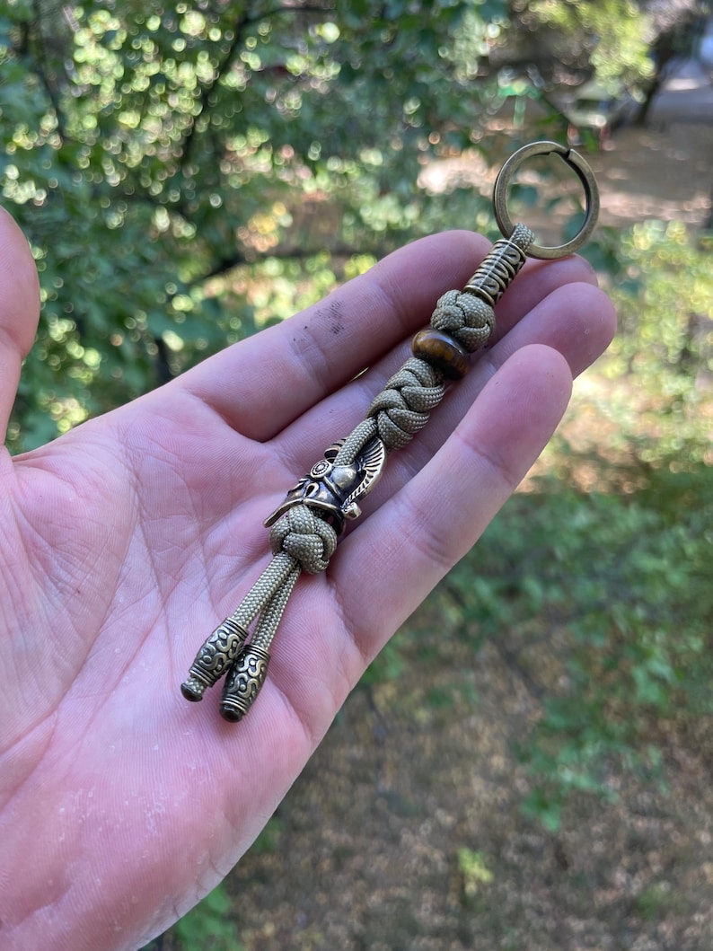 K&ouml;nnte beinhalten: Ein gr&uuml;ner Paracord-Schl&uuml;sselanh&auml;nger mit einem Wikingerhelm-Anh&auml;nger aus Messing und einer braunen Holzperle. Der Schl&uuml;sselanh&auml;nger hat zwei Messing-Endkappen mit einem dekorativen Muster.