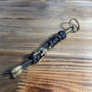 K&ouml;nnte beinhalten: Ein schwarzer Paracord-Schl&uuml;sselanh&auml;nger mit einem Messing-Schl&uuml;sselring und einer Messingperle. Der Schl&uuml;sselanh&auml;nger hat einen dekorativen Messing-Anh&auml;nger mit einem Knoten oben.