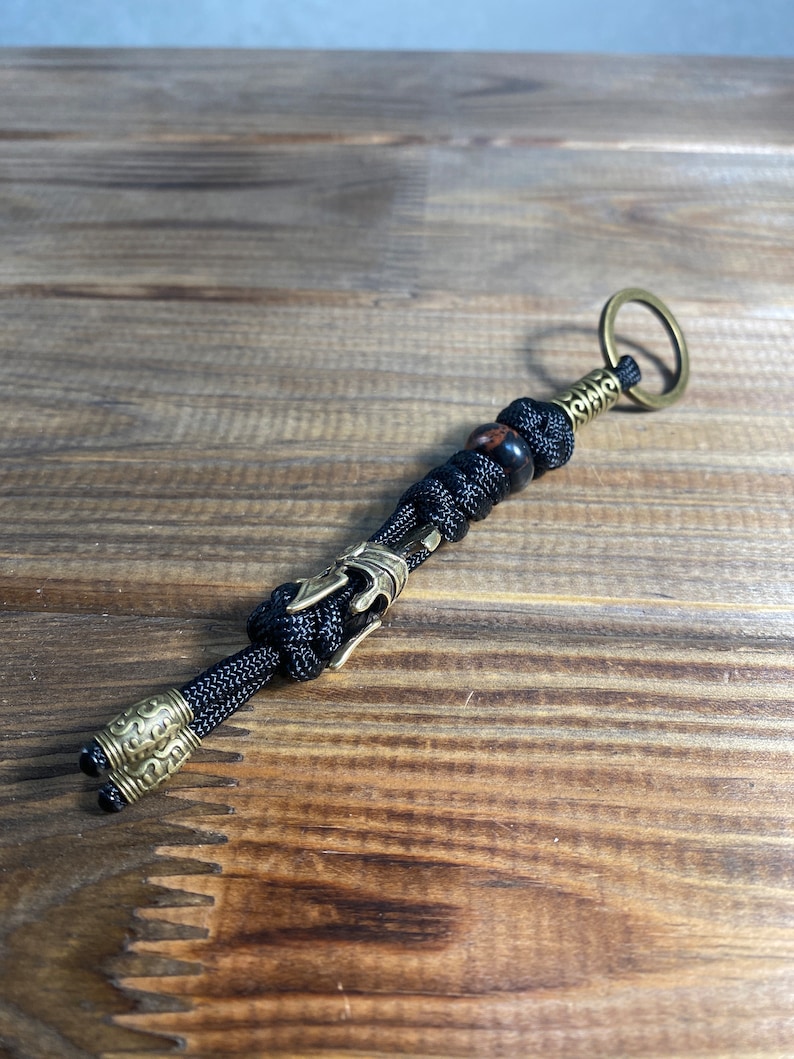K&ouml;nnte beinhalten: Ein schwarzer Paracord-Schl&uuml;sselanh&auml;nger mit einem Messing-Schl&uuml;sselring und einer Messingperle. Der Schl&uuml;sselanh&auml;nger hat einen dekorativen Messing-Anh&auml;nger mit einem Knoten oben.