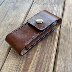 May include: A brown leather sheath with a brass snap closure. The sheath is rectangular and appears to be hand-stitched. The leather has a rich, dark brown color and a slightly textured surface. The sheath is designed to hold a small tool or knife.