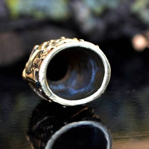 May include: A brass thimble with an intricate design. The thimble is round and has a small opening at the top. The thimble is sitting on a black surface.
