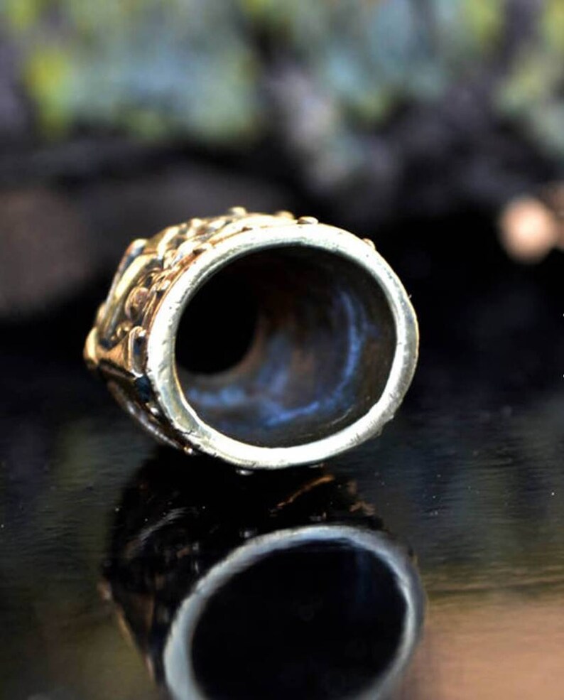 May include: A brass thimble with an intricate design. The thimble is round and has a small opening at the top. The thimble is sitting on a black surface.
