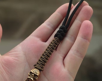 May include: A brown and black paracord keychain with a gold skull charm. The skull has intricate details and a Celtic knot design.