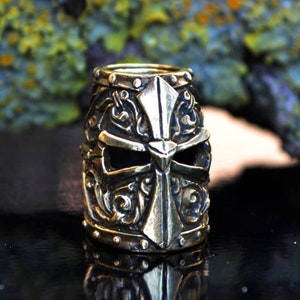 May include: A detailed brass bead in the shape of a knight's helmet with a cross design. The helmet has intricate details and a dark eye opening.