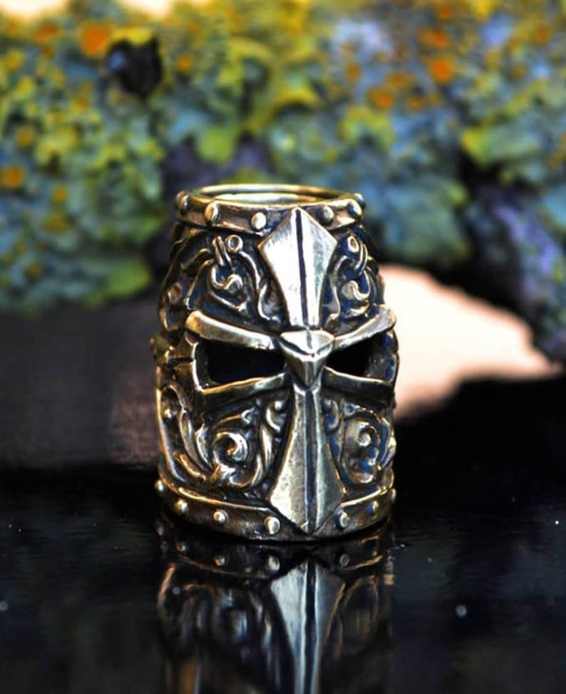 May include: A detailed brass bead in the shape of a knight's helmet with a cross design. The helmet has intricate details and a dark eye opening.
