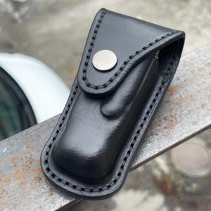 Puede incluir: Una funda de cuero negro con una solapa asegurada por un broche plateado. La funda tiene un borde cosido y parece estar diseñada para guardar una herramienta pequeña o un cuchillo. El cuero tiene un acabado brillante.