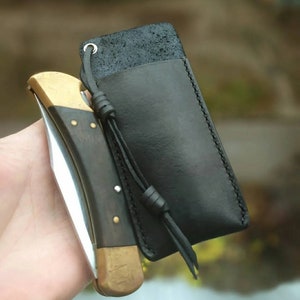 Peut inclure: Un étui à couteau en cuir noir avec un couteau à manche en bois brun et un accent en laiton. L'étui est doté d'une sangle en cuir noir avec un nœud.