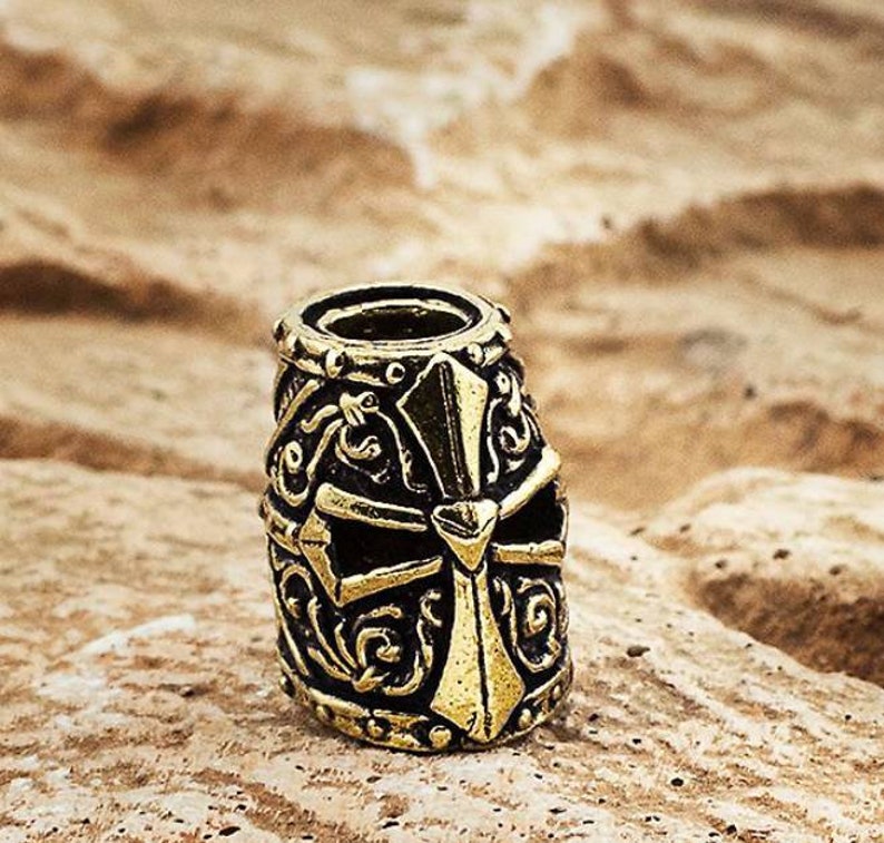 May include: A gold-coloured metal bead with an intricate design featuring a cross and floral patterns. The bead is cylindrical in shape and has a smooth, polished finish.
