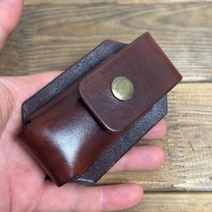 May include: A dark brown leather pouch with a smooth finish and a rectangular flap secured by a single round brass-coloured metal snap button. The pouch features a structured design with a reinforced back panel and visible stitching along the edges.