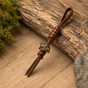 Op de afbeelding: Een bruin leren sleutelhanger met een bronzen kikkerbedel. De sleutelhanger heeft een lus aan de bovenkant en een gespleten uiteinde. De kikkerbedel is gedetailleerd en voegt een unieke touch toe. Het leer lijkt goed gemaakt.