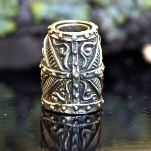 May include: A silver-toned metal bead with intricate, detailed carvings. The bead has a cylindrical shape with a rounded top and bottom. The bead is decorated with a geometric pattern and floral motifs.