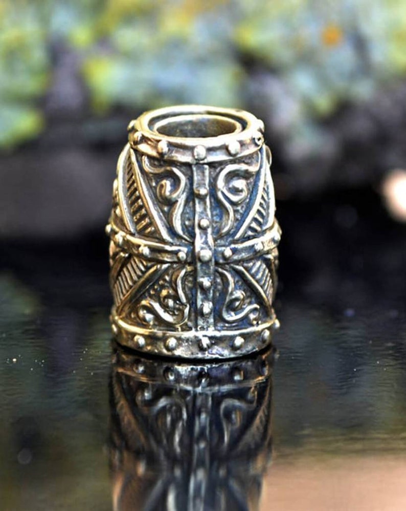 May include: A silver-toned metal bead with intricate, detailed carvings. The bead has a cylindrical shape with a rounded top and bottom. The bead is decorated with a geometric pattern and floral motifs.