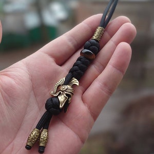 K&ouml;nnte beinhalten: Ein schwarzer Paracord-Schl&uuml;sselanh&auml;nger mit einem goldfarbenen r&ouml;mischen Helm-Anh&auml;nger und einer Tigerauge-Perle.