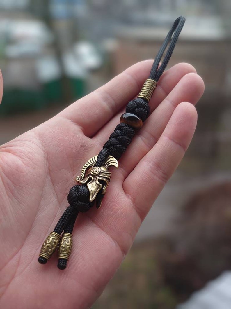 K&ouml;nnte beinhalten: Ein schwarzer Paracord-Schl&uuml;sselanh&auml;nger mit einem goldfarbenen r&ouml;mischen Helm-Anh&auml;nger und einer Tigerauge-Perle.