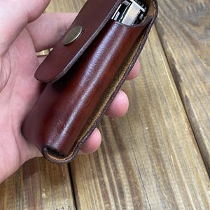 Pode incluir: Uma capa de couro castanho com fecho de pressão, contendo uma multi-ferramenta prateada e preta. A capa tem um acabamento liso e brilhante e um botão de pressão cor de latão. A multi-ferramenta é parcialmente visível.